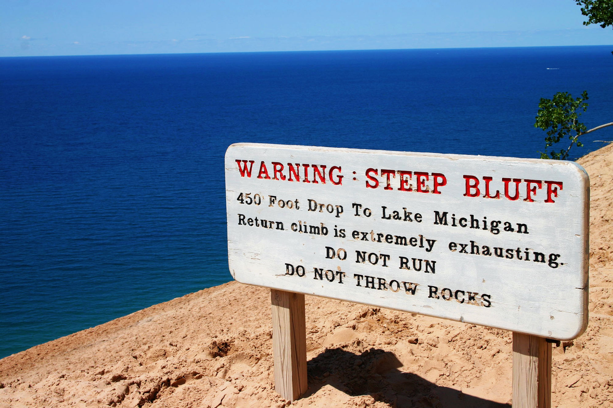 A white sign with red and black text warning guests about the terrain of sand and water behind the sign.